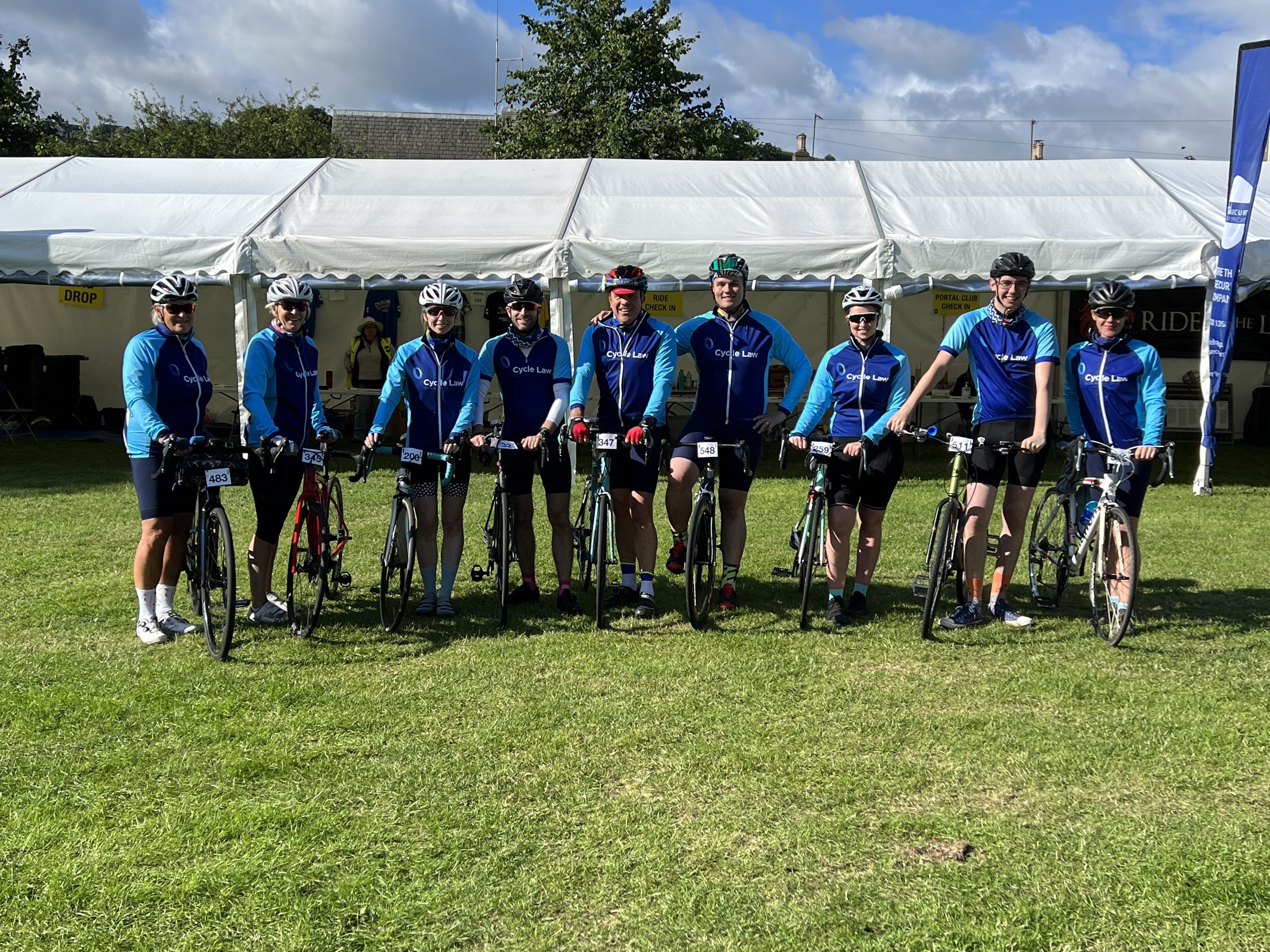 The photo show the Cycle Law Scotland legal team dresses in their cycle team kit, with their bikes at an event. There is a large marquee in the background