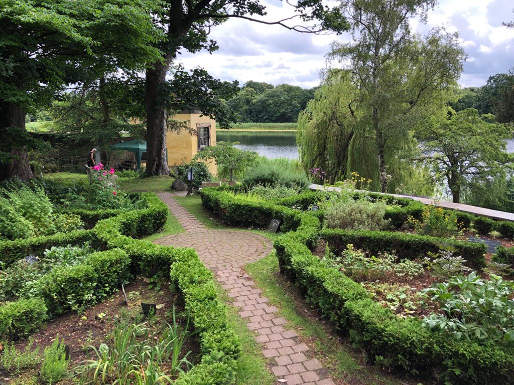 Photo shows part of Dr Neil's Garden. A red brick path in the centre of the photo leads between low box hedges, surrounding flower boarders and trees. At the end of the path is a low sandstone building and a view of the loch.