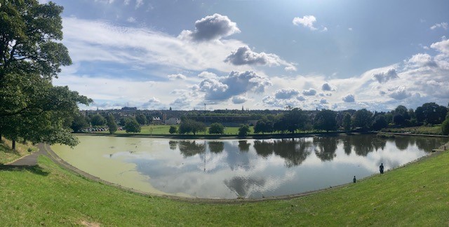 Photo taken with a wide-angle lens, showing park with grass in the foreground, with trees around it. In the middle is a lake which is reflecting a partly clouded sky,