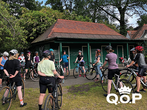 Image shows a group of stationary cyclists, gathered in a circle in front of a hut